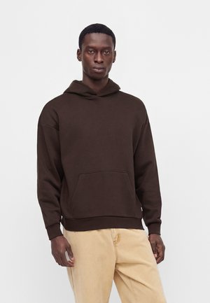 Junger Mann trägt einen dunkelbraunen Hoodie und hellbeige Hose und steht vor einem schlichten weißen Hintergrund.