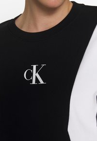 Sudadera negra con panel lateral blanco, con un prominente logo "cK" en blanco en el pecho. Tela suave con escote redondo.