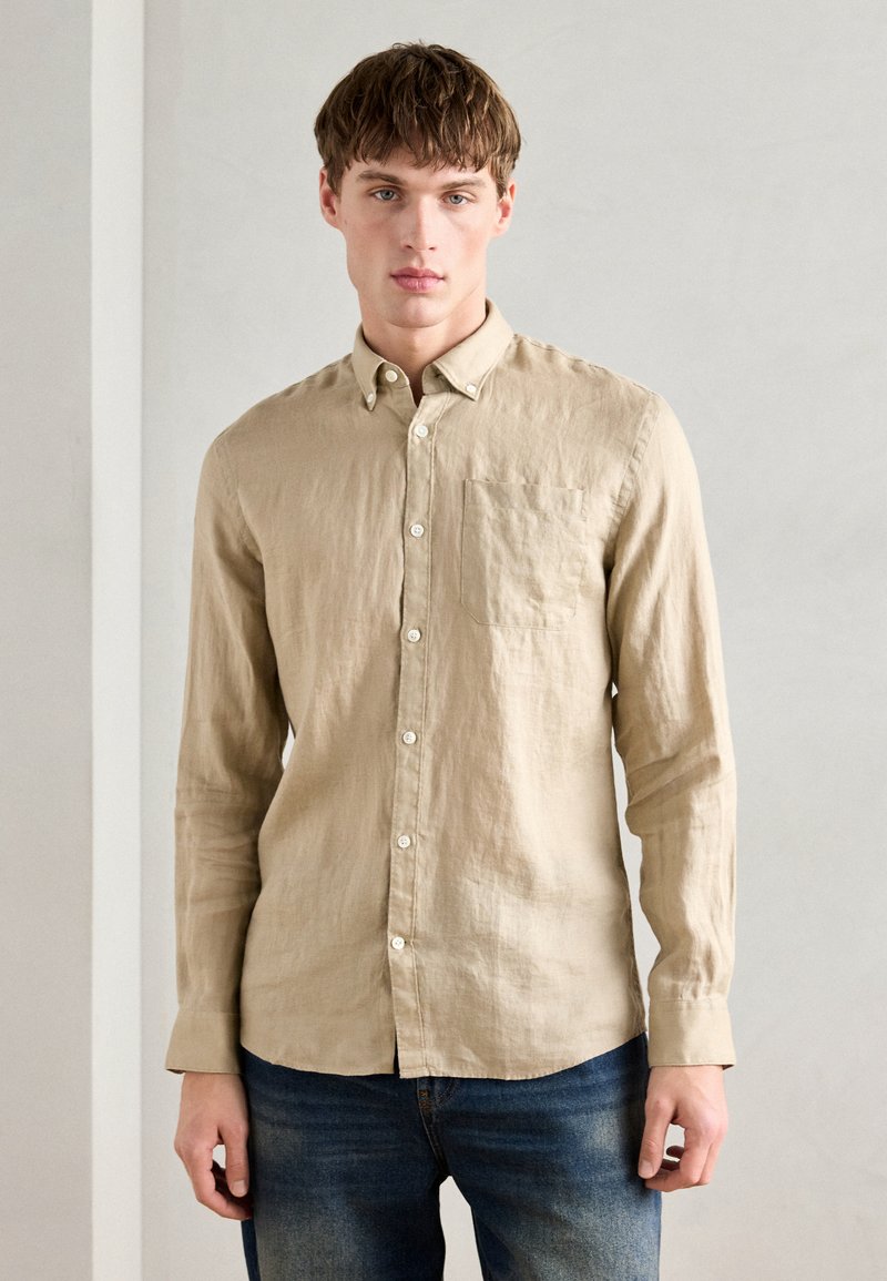 Camisa bege de mangas longas, com botões, feita de linho leve; possui um bolso no peito, botões brancos e um corte casual com a barra reta.