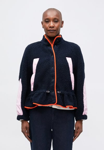 Veste en polaire marine avec des panneaux latéraux roses, des bordures orange et un ourlet peplum. Présente un col haut et une fermeture à boutons-pression. Surface texturée.