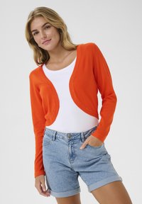 Cardigan corto arancione realizzato in tessuto a maglia leggero, con un davanti arrotondato e una consistenza morbida, abbinato a una t-shirt bianca e a pantaloni shorts in denim.