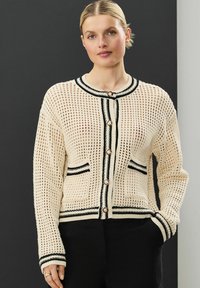 Beżowy sweter z dzianiny o otwartej strukturze, z czarnymi wykończeniami i złotymi guzikami. Posiada dwie przednie kieszenie i długie rękawy.