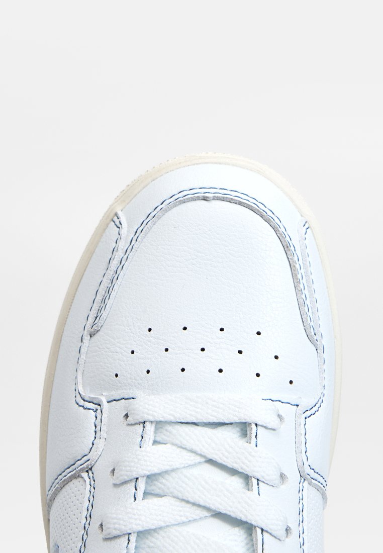 Zapatilla de cuero blanca con puntera redonda, detalles perforados y costuras claras. Cuenta con cordones planos y una suela de goma texturada en color blanco roto.