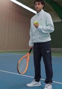 Casaco desportivo branco com fecho éclair e gola alta. Calças de treino azuis. Segurando uma bola de ténis amarela e uma raquete de ténis laranja num campo azul.