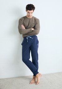 Brun langermet skjorte med logo, kombinert med marineblå joggebukser med snøring i livet, på en barføtt modell mot en lys vegg.