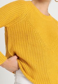 Gelber Pullover aus Strick mit strukturiertem Muster, runder Ausschnitt und gerippten Bündchen, in lockerer Passform und mit sichtbaren seitlichen Schlitzen.
