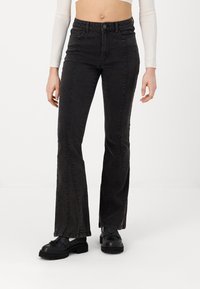 Jeans flare em denim preto com cintura alta, apresentando costura de contraste e costuras laterais. Combinados com sapatos pretos chunky.