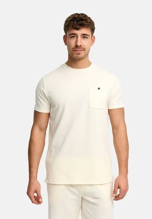 Ung mand med kort krøllet hår iført tekstureret off-white kortærmet skjorte og matchende bukser, stående mod ensfarvet baggrund.