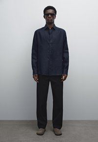 Black linen long-sleeve shirt, buttoned front, paired with black loose trousers and gray slip-on shoes. Model wearing dark sunglasses.