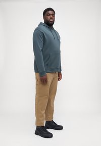 Sudadera verde azulado con bolsillo frontal, combinada con pantalones cargo color beige y botas negras con cordones, caracterizada por un acabado texturizado y un diseño deportivo.