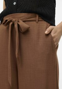 Pantalon marron taille haute avec ceinture en tissu nouée, main dans la poche, associé à un haut en tricot noir avec détail de boutons.
