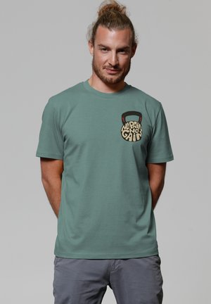 T-shirt en coton vert clair avec un col rond et des manches courtes. Présente un graphique de kettlebell avec le texte "No Pain No Gain" sur la poche poitrine.