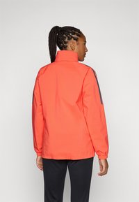 Koraalkleurig jack met hoge kraag en elastische manchetten, met blauwe strepen langs de schouders. Glad textuur, lichtgewicht materiaal.