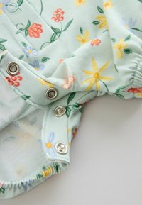 Gros plan sur un tissu vert menthe à imprimé floral jaune, rouge et bleu avec trois boutons-pression argentés et des bords élastiques.