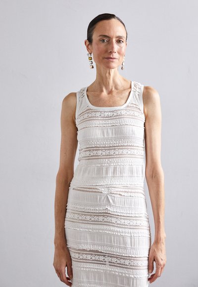 Robe blanche sans manches en tissu texturé, avec des motifs en dentelle et des rayures horizontales. Le modèle porte des boucles d'oreilles audacieuses.
