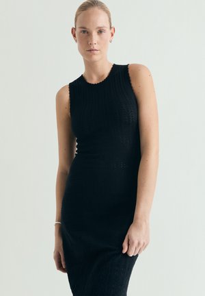 Mujer con el pelo recogido, vestida con un vestido negro sin mangas y ajustado de punto, de pie contra un fondo claro y liso.