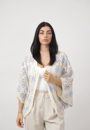 Vrouw met lang zwart haar, gekleed in een wit shirt, beige broek en een losse gehaakte cardigan met blauwe en crèmekleurige patronen.