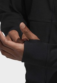 Svart sportjacka med detaljer som visar den texturerade tyget på ärmarna, tumhål och en åtsittande design som framhäver händer som greppar manschetterna.