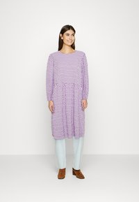 Vestido longo roxo de mangas compridas com um padrão de bolinhas, cintura marcada e corte descontraído. Combinado com jeans azul claro e botas castanhas de tornozelo.