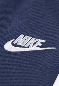 Jūros mėlynos spalvos audinys su minkšta tekstūra, kuriame išryškėja baltas išsiuvinėtas Nike logotipas, rodantis ryškų šriftą ir sklandžias linijas.