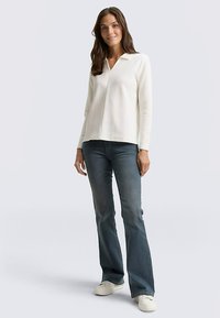 Długi rękaw, bluzka z dekoltem w serek z kremowej dzianiny, zestawiona z niebieskimi dzwonowatymi dżinsami i białymi trampkami. Gładka faktura, casualowy styl.