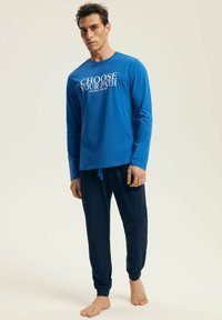 Man stands barefoot wearing blue long-sleeve shirt with "CHOOSE YOUR PATH" text and dark blue jogger pants against a plain light background.