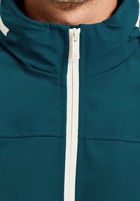 Tealfärgad jacka med hög krage, med en vit dragkedja och kontrasterande vita ränder längs kragen. Slät textur med synliga sömmar.