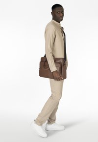 Braune Leder-Umhängetasche mit glänzender Oberfläche, ausgestattet mit einer Fronttasche und einem abnehmbaren Schultergurt, gehalten von einer Person in hellbeiger Kleidung.