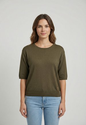 Mujer con cabello castaño hasta los hombros, vistiendo un suéter de manga corta color verde oliva y jeans azul claro, de pie frente a un fondo sencillo.