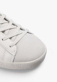 Zapatillas de deporte en gris claro con parte superior de ante y cuero, suela de goma texturizada y cordones planos con un acabado brillante. Diseño de punta redondeada.