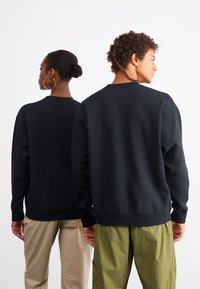 Must tume fliisist, millel on ümar kaelus, soonikkoes varrukad ja serv. Kaks figuuri, üks kantud beežide pükstega ja teine roheliste pükstega.