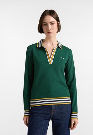 Donna che indossa un polo verde scuro a maniche lunghe con colletto, polsini e orlo a righe, abbinato a jeans blu, in piedi davanti a uno sfondo semplice.