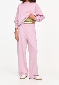 Svetle růžová mikina s žebrovaným výstřihem, spárovaná s odpovídajícími kalhotami s širokými nohavicemi. Oblečení obsahuje zelený top jako spodní vrstvu.