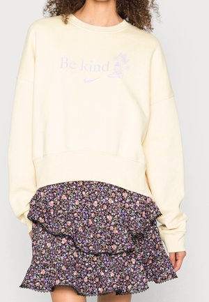Person iført en cremefarvet sweatshirt med teksten "Be kind" og blomsterprint, kombineret med en sort blomsterprintet flæse-nederdel.
