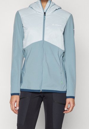 Persoon draagt een lichtblauw en bleekblauw gewatteerd jack met capuchon met ritssluiting aan de voorkant en zijzakken, gecombineerd met een zwarte broek.