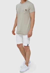 Mężczyzna w beżowej koszulce z krótkim rękawem z paskowanym kieszonką na piersi, białych szortach podwiniętych na nogawkach i białych trampkach, stojący na szarym tle.