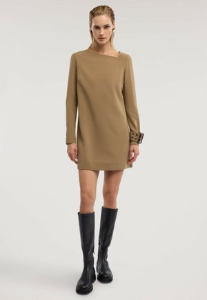 Femme portant une mini-robe marron à manches longues avec un décolleté asymétrique et des bottes noires montant jusqu'aux genoux, debout sur un fond uni.
