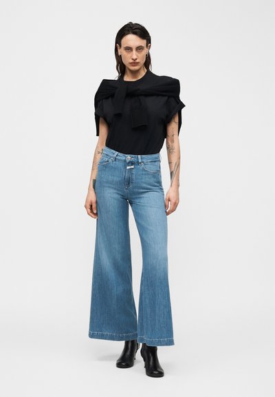 Νεαρή γυναίκα με τατουάζ φοράει μαύρο τοπ, μαύρο πουλόβερ περασμένο στους ώμους, φαρδιά μπλε τζιν παντελόνια και μαύρα μποτάκια αστραγάλου σε λευκό φόντο.