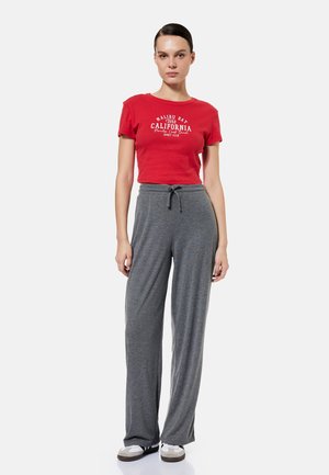 Kobieta stojąca, ubrana w czerwoną krótką koszulkę z napisem „California” oraz szare, szerokie spodnie z sznurkiem, białe trampki na białym tle.