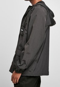 Black lightweight jacket with a zip closure, hood, and elastic cuffs. Features a front pocket with a branded patch for added detail.