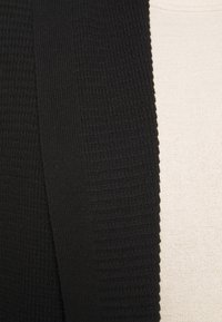 Schwarze Strickjacke mit strukturiertem Muster, offener Vorderseite und langen Ärmeln, neben einem hellbeigen Oberteil präsentiert.