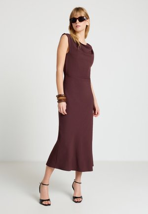 Femme portant une robe midi sans manches marron foncé, des sandales à talons hauts noires, de grandes lunettes de soleil et des bracelets en bois, debout devant un fond uni.