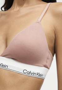 Γυναίκα φοράει ένα ανοιχτό ροζ bralette Calvin Klein με λεπτές τιράντες και λευκή ελαστική ταινία με μαύρο λογότυπο.
