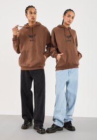 Felpe marroni con grafiche nere, abbinate a jeans larghi neri e blu chiaro. Entrambi indossano scarpe con piattaforma nere.