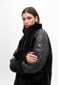 Black plush jacket with a glossy fabric finish. Features a high collar, padded sleeves, and an emblem on the left arm. Smooth texture.