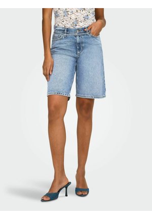 Žena oblečená v stredne modrých denimových bermudách, kvetinovom čipkovanom tielku bez rukávov a modrých otvorených lodičkách s podpätkom, stojí s jednou rukou v vrecku.
