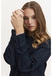 Cămașă din denim închis cu manșete cu butoni, guler și detalii cu cusături deschise. Modelul are părul ondulat și își ține mâinile împreunate.