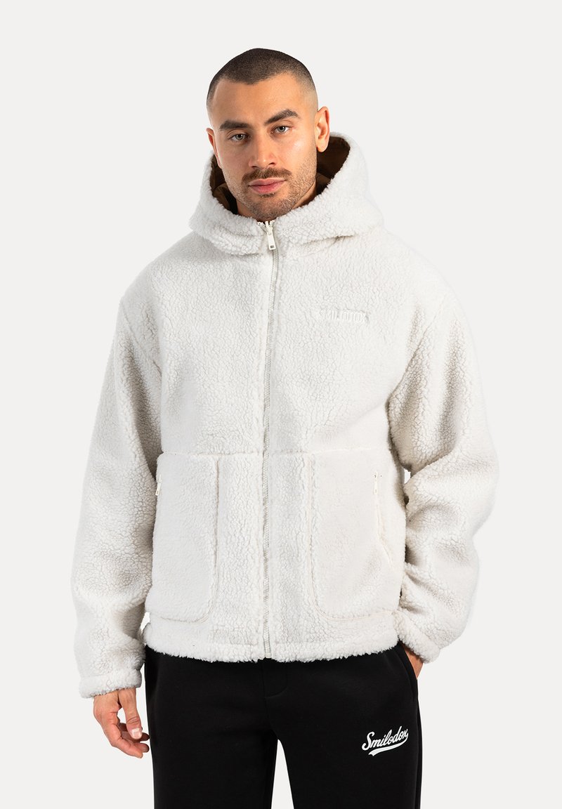 Sudadera blanca de forro polar con cierre de cremallera, grandes bolsillos frontales y un cuello alto. Textura suave, ajuste relajado, incluye una capucha forrada.