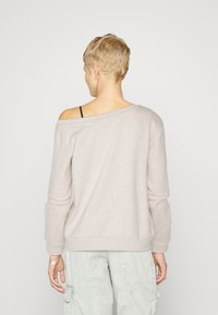 Helle beige schulterfreie Sweatshirt mit langen Ärmeln, geripptem Saum und weicher Textur. Über einem schwarzen Trägershirt getragen, kombiniert mit hellen Hosen.
