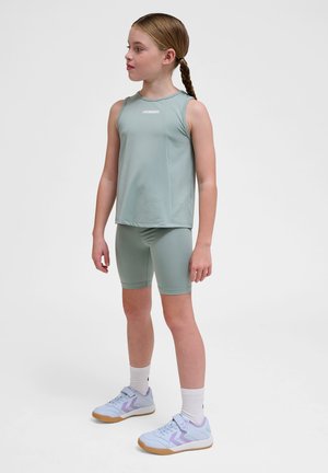 Ung pige med flettet hår iført en lysegrøn ærmeløs top, matchende cykelshorts, hvide sokker og lyse lilla sneakers, stående sideværts.